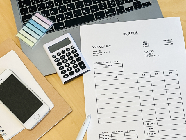 机の上に置かれた御見積書の用紙。電卓、ノートパソコン、スマートフォンが並んでいる
